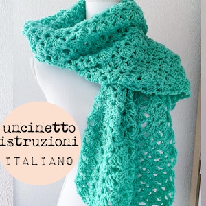 May include: A teal crochet scarf with a lacy pattern. The scarf is draped over a white mannequin. The text "uncinetto istruzioni ITALIANO" is printed on a peach-colored circle.