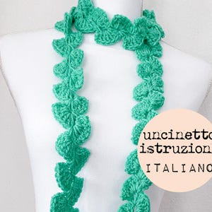 May include: A green crocheted scarf with a leaf pattern. The scarf is displayed on a white surface with the text "uncinetto istruzioni ITALIANO" in a circle.