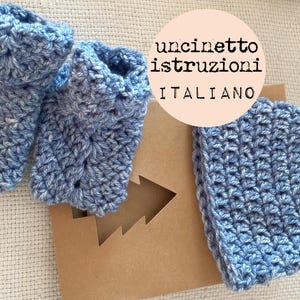 May include: A set of blue crocheted baby booties and a blue crocheted baby hat. The hat and booties are on a brown background with a white paper cutout of a Christmas tree. The background also has a white circle with the text "uncinetto istruzioni ITALIANO".
