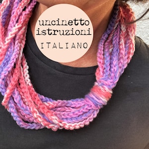 May include: A close-up of a pink and purple crocheted necklace. The necklace is made of multiple strands of yarn and is worn by a person. The text "uncinetto istruzioni italiano" is visible on a white circle.