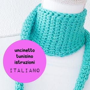 May include: A turquoise crocheted scarf is displayed on a white mannequin head. The scarf is made with a textured stitch and has long, dangling ends. A pink circle with Italian text is in the lower left corner.
