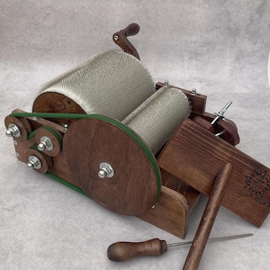 May include: A wooden drum carder featuring two large cylinders covered in fine metal teeth, a green drive belt, and a hand crank. The device includes a wooden feed tray with a sun engraving, a wooden doffer stick, and a wooden cleaning tool.