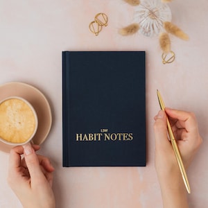 May include: A navy blue notebook with the gold text "HABIT NOTES". A gold pen is positioned to the right. A cup of coffee and gold paper clips are also visible on a pale pink surface.