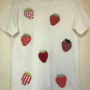 May include: White t-shirt decorated with appliqued strawberries in diverse patterns and colours. The strawberries have red bodies and green leafy tops. The shirt is hanging on a black hanger.