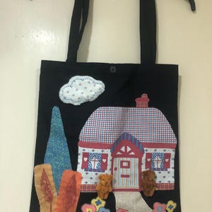 May include: Black tote bag with a charming house design. The house has a red and white checked roof, a red door, and two small teddy bears. Appliqued details include a cloud, trees, and flowers.