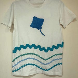 Op de afbeelding: Wit T-shirt met een blauwe rog en golvende lijnen in blauw en groen. Het shirt met korte mouwen heeft een ronde hals. Het ontwerp is gedrukt op de voorkant van het shirt, dat is gemaakt van een zachte, lichte stof.