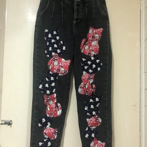 Könnte beinhalten: Dunkelgraue Jeans mit hoher Taille und Knopfverschluss. Die Jeans sind mit applizierten Katzenmotiven in rot-weißen Mustern auf marineblauem Hintergrund verziert. Die Katzen sind in vertikalen Reihen entlang der Beine angeordnet.