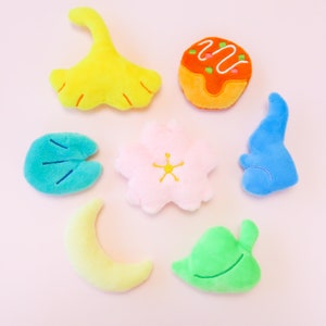 Puede incluir: Siete juguetes de peluche de diferentes formas y colores, incluyendo una hoja de ginkgo amarilla, una flor rosa, un pez azul, una hoja verde, una luna creciente amarilla, un nen&uacute;far azul y un takoyaki naranja.