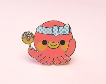 Puede incluir: Pin esmaltado de un pulpo rosa con contorno dorado. El pulpo lleva una diadema y un lazo de lunares azules y blancos. Sostiene un pincho de takoyaki. El pin tiene el texto "tokyo shop".