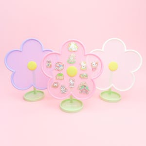 May include: Three pink flower-shaped pin holders with mesh netting. The center holder has a variety of colorful enamel pins with cartoon characters and designs.