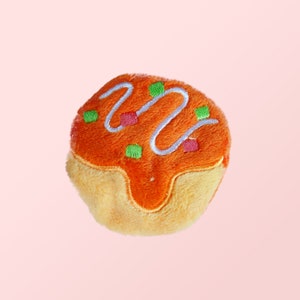 Puede incluir: Un juguete de peluche con forma de donut con glaseado naranja y glaseado blanco. El donut tiene chispas verdes y rosas.
