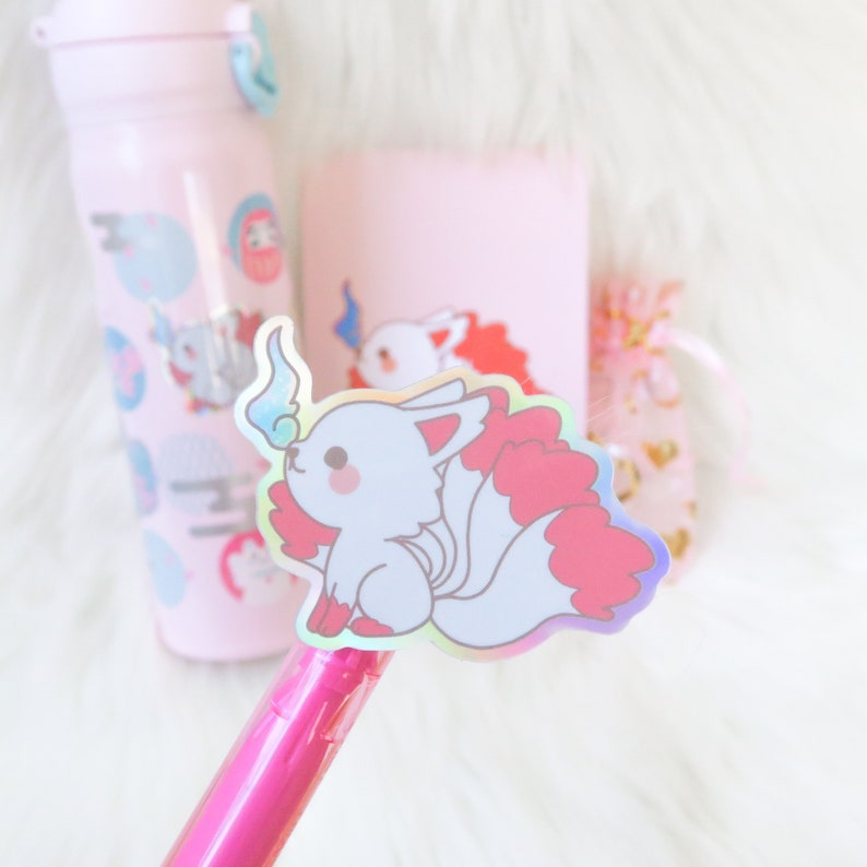 May include: A holographic sticker of a white fox with red fur and a blue flame on its tail. The sticker is on a pink pen.