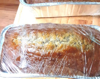 May include: Two loaves of freshly baked banana bread in aluminum foil pans, wrapped in clear plastic wrap. The bread has a golden-brown crust and is sitting on a wooden surface.