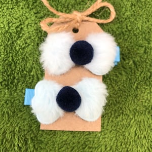 May include: Two white fluffy hair bows with blue ribbon clips and black pom-poms. The bows are attached to a brown card with twine.