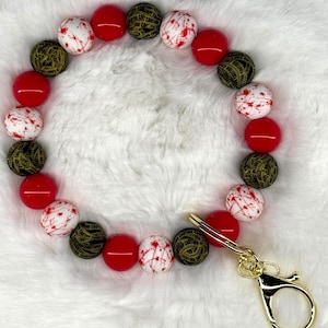 May include: A beaded wristlet featuring a variety of colorful beads. The wristlet includes red, white with red splatter, and black and gold patterned beads. A gold-colored clasp is attached to the wristlet.