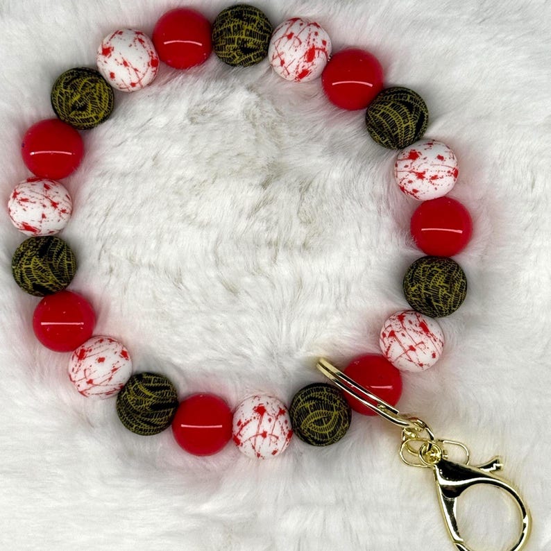 May include: A beaded wristlet featuring a variety of colorful beads. The wristlet includes red, white with red splatter, and black and gold patterned beads. A gold-colored clasp is attached to the wristlet.