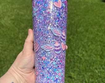 May include: A tall, glitter-filled tumbler with a black lid. The tumbler is covered in purple, blue, and pink glitter. It features bee and heart decals, along with the word "believe" in cursive. Floral and star accents complete the design.