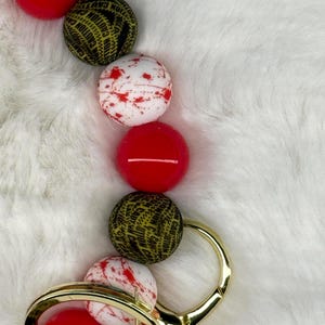 May include: A gold-colored keychain with a series of colorful beads. The beads include red, white with red splatter, and black with yellow "CAUTION" text. The keychain is on a white, fluffy surface.