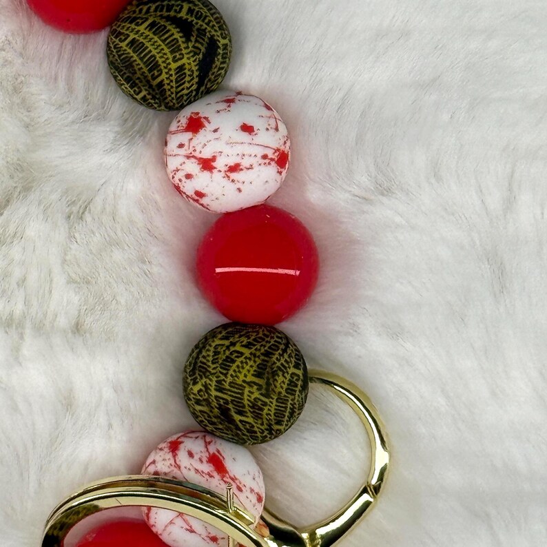 May include: A gold-colored keychain with a series of colorful beads. The beads include red, white with red splatter, and black with yellow "CAUTION" text. The keychain is on a white, fluffy surface.