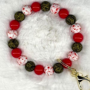 May include: A beaded keychain bracelet featuring red, white, and olive green beads. The white beads have red splatter designs. The bracelet is attached to a gold-colored clasp.