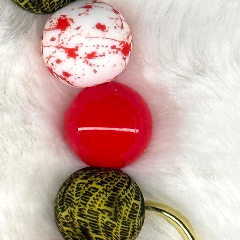 May include: Close-up of a necklace featuring large, round beads. The beads are white with red splatters, solid red, and yellow with black text that reads "CAUTION". A gold-colored clasp is visible.