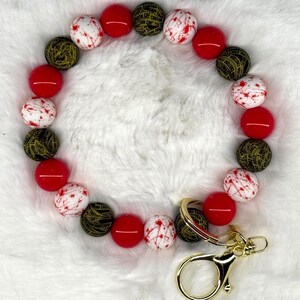 May include: A beaded keychain wristlet featuring a mix of red, white with red splatter, and black and gold patterned beads. The wristlet has a gold-colored clasp and key ring.