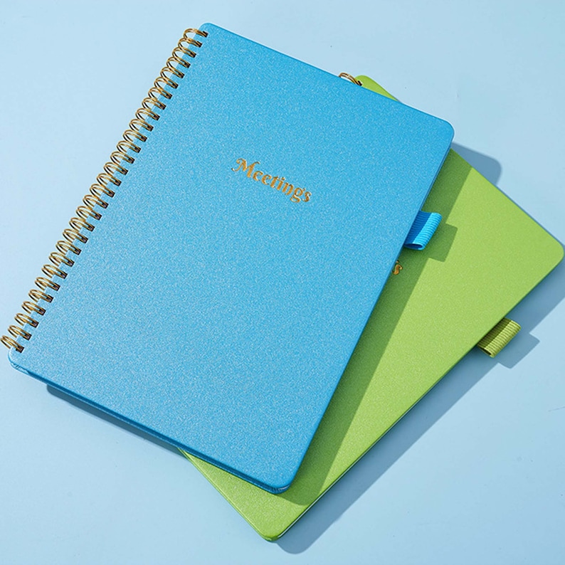 May include: Two spiral bound notebooks, one blue and one green. The blue notebook has the word "Meetings" written in gold on the front cover.