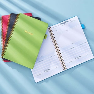 May include: A stack of three green, black, and red spiral bound notebooks with gold coils. The top notebook is open to reveal a blue page with the text "Meeting Notes" and a table for recording meeting details.