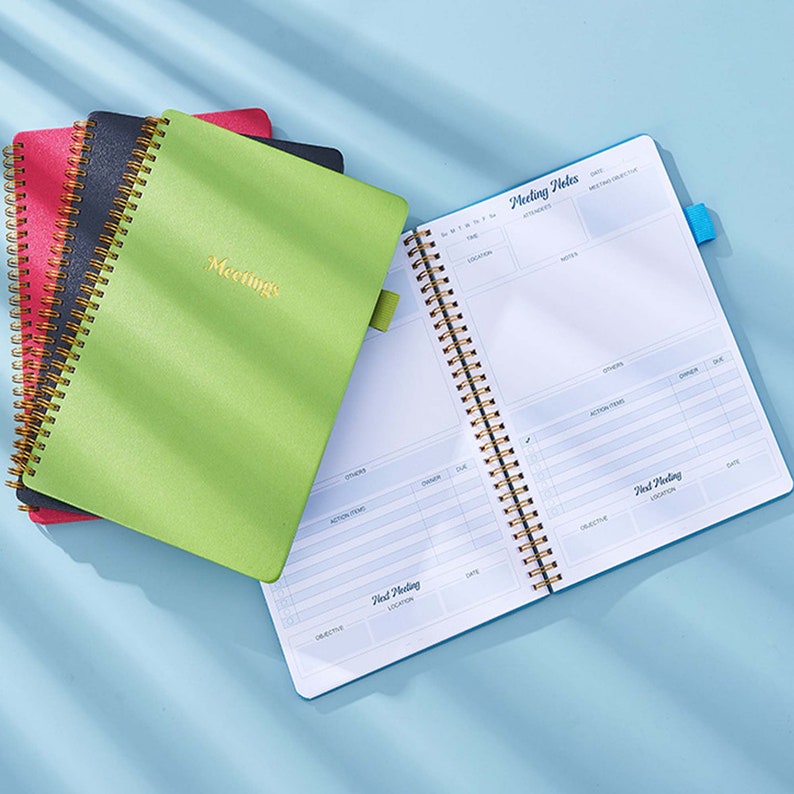 May include: A stack of three green, black, and red spiral bound notebooks with gold coils. The top notebook is open to reveal a blue page with the text "Meeting Notes" and a table for recording meeting details.