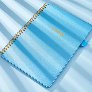 May include: A light blue spiral bound notebook with a gold coil and a gold elastic band. The cover has the word "Meetings" printed in gold lettering.