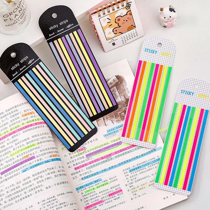 May include: Two packs of colorful sticky notes. The first pack has pastel colored strips with the text "sticky strips book - home - office". The second pack has neon colored strips with the text "sticky notes".