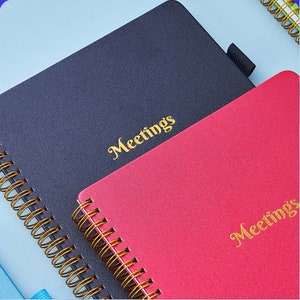 May include: Two spiral bound notebooks with gold wire binding. The notebook on the left is black with the word "Meetings" in gold lettering. The notebook on the right is red with the word "Meetings" in gold lettering.