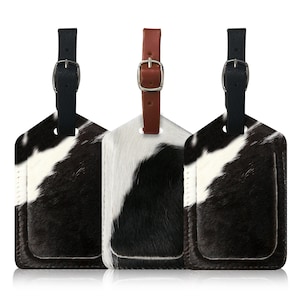 Puede incluir: Tres etiquetas de equipaje con un patrón de piel de vaca en blanco y negro. Cada etiqueta tiene una correa de cuero con una hebilla plateada. Una correa es marrón y las otras dos son negras. Las etiquetas tienen forma rectangular con la parte inferior redondeada.