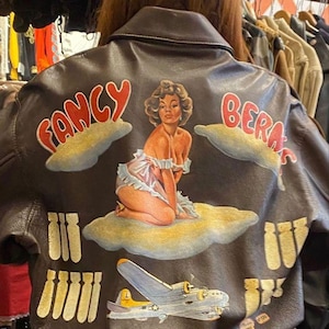 May include: A brown leather jacket with a painted design on the back. The design includes the words "Fancy Beras", a pin-up girl, a B-17 bomber, and stylized bombs. The jacket has a classic bomber style.