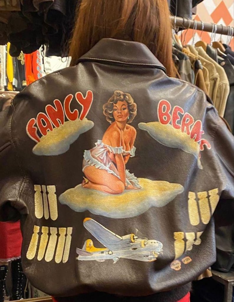 May include: A brown leather jacket with a painted design on the back. The design includes the words "Fancy Beras", a pin-up girl, a B-17 bomber, and stylized bombs. The jacket has a classic bomber style.