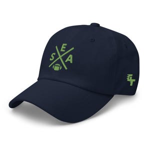 May include: Navy blue baseball cap with a curved brim. The cap features the word "SEA" in a green, stylized design with a football in the center. A green logo is on the side.