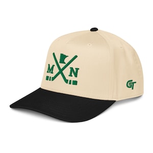 Puede incluir: Una gorra de béisbol de color crema con visera negra y un botón negro en la parte superior. El frente presenta un diseño bordado en verde de dos palos de hockey cruzados, las letras M y N, y el contorno del estado de Minnesota. Las letras GT están bordadas en verde en el lateral.