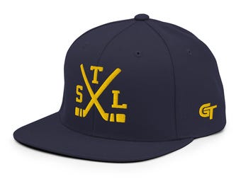 May include: A navy blue baseball cap with a flat brim. The front features yellow embroidered text reading STL arranged around two crossed hockey sticks. The side of the cap displays a yellow embroidered GT logo. The cap has a structured crown.