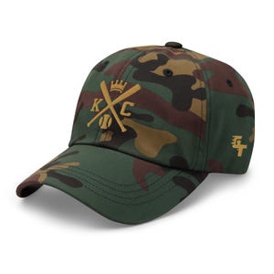 May include: A camouflage baseball cap with a gold embroidered logo featuring a crown, crossed baseball bats, and the letters "KC".