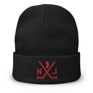 Könnte beinhalten: Schwarze Mütze mit einem roten Stickmotiv aus gekreuzten Hockeyschlägern, dem Bundesstaat New Jersey und den Buchstaben "NJ".