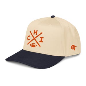 May include: A cream-colored baseball cap with a dark navy blue brim and a dark navy blue button on top. The front features an embroidered orange design with crossed lines, the letters C, H, and I, and a football. The letters GT are on the side.