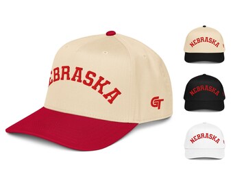 May include: A baseball cap with a cream crown, a red brim, and a red button on top. The word NEBRASKA is embroidered in red across the front in an arched font. A red GT logo is on the side. Three additional caps are shown in the background.