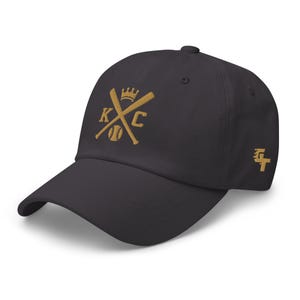 May include: A dark gray baseball cap with a gold embroidered design featuring crossed baseball bats, a baseball, and the letters "KC" with a crown above the letters.