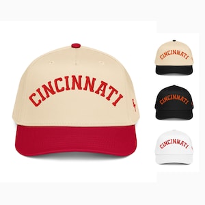May include: A baseball cap with a cream-coloured crown and a red brim. The word "CINCINNATI" is arched across the front in red lettering. Additional caps in black, white, and cream are also shown.