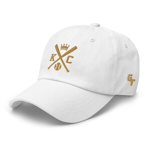 May include: White baseball cap with a gold embroidered design featuring crossed baseball bats, a baseball, and the letters "KC" with a crown above the bats.