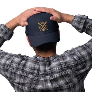 May include: A navy blue baseball cap with a gold embroidered design featuring a crown, crossed baseball bats, and the letters "KC".