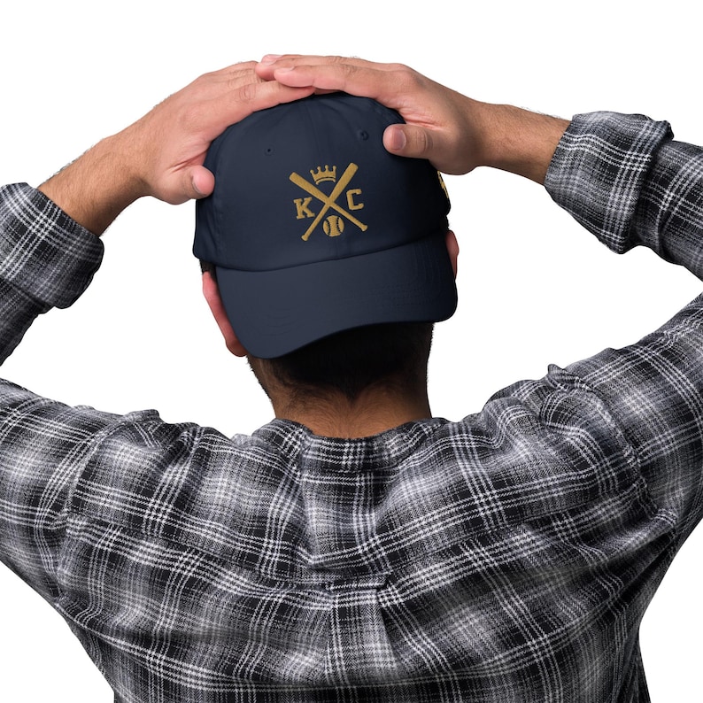 May include: A navy blue baseball cap with a gold embroidered design featuring a crown, crossed baseball bats, and the letters "KC".