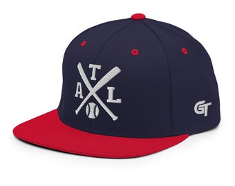 Könnte beinhalten: Eine marineblaue Baseballkappe mit rotem flachem Schirm und roten Ösen. Die Vorderseite zeigt eine weiße Stickerei aus zwei gekreuzten Baseballschlägern, einem Baseball und den Buchstaben ATL. Die Seite zeigt die weißen Buchstaben GT.