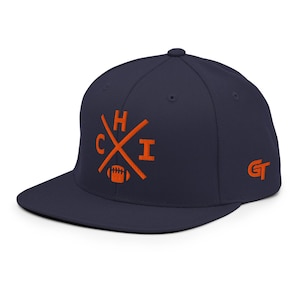 Könnte beinhalten: Eine marineblaue Snapback-Cap mit einem gestickten orangefarbenen Design auf der Vorderseite, das die Buchstaben C, H und I um gekreuzte Linien über einem Football-Symbol zeigt. An der Seite befindet sich ein gesticktes orangefarbenes GT-Logo.
