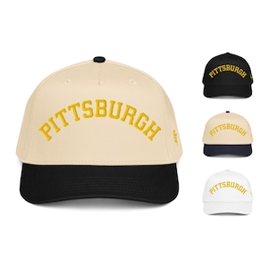 以下が含まれることがあります： 前面に黄色でPITTSBURGHの刺繍が施されたベースボールキャップ。メインのキャップはクリーム色で、黒いツバと黒いボタンが上部に付いています。右側には、黒、黒いツバのクリーム色、白の3つのバリエーションが追加で表示されています。